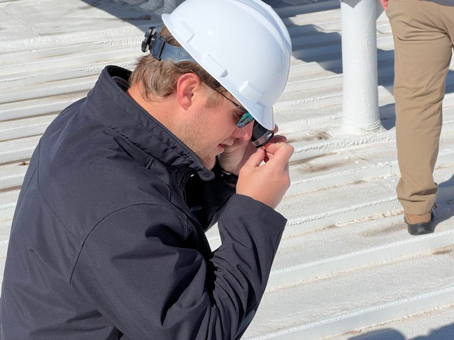 Construction worker in a white hard hat and safety glasses adjusting his eyewear on a metal rooftop surface.